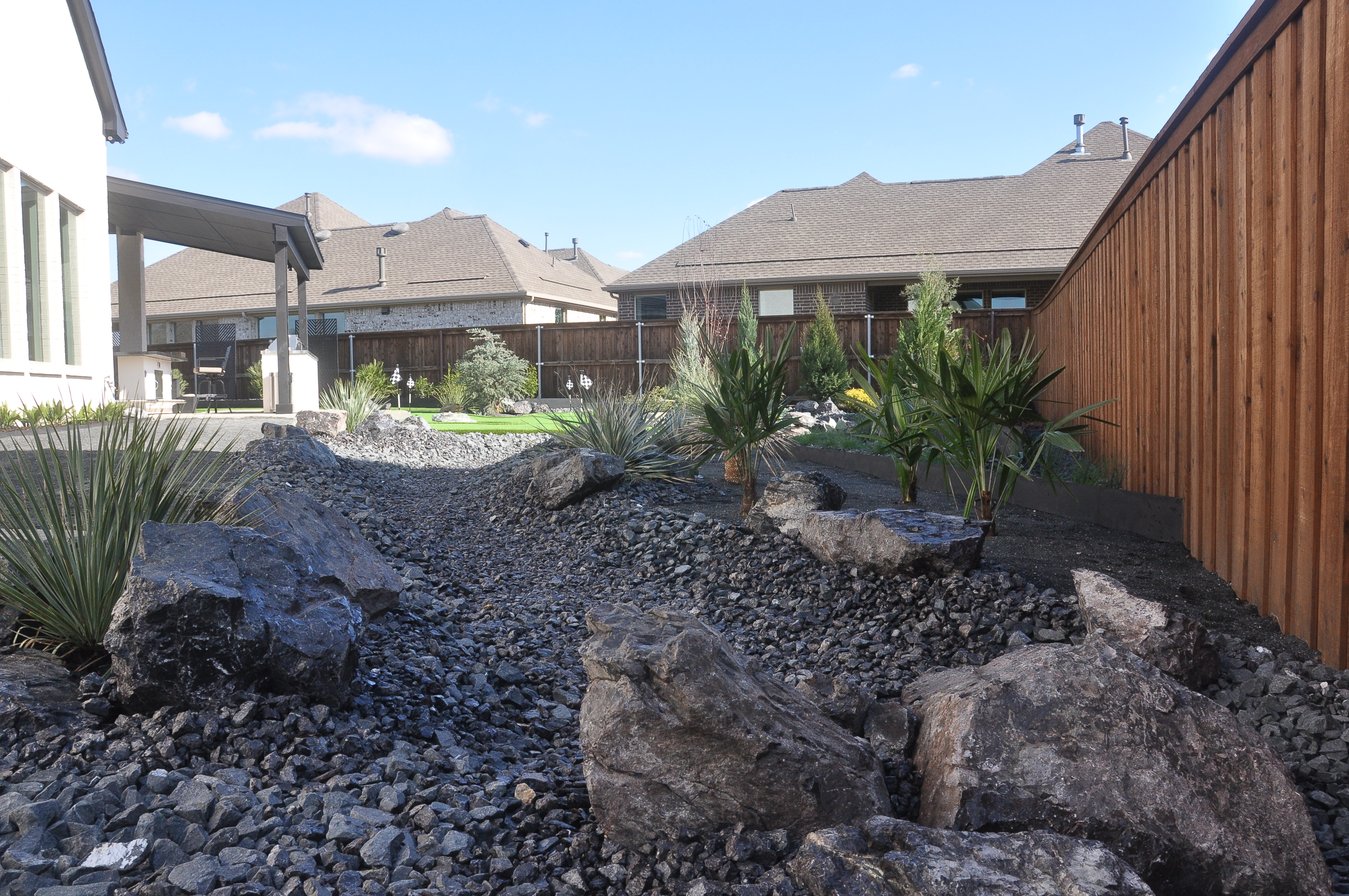 A residential backyard showcases a dry creek bed drainage solution constructed with dark gravel and large boulders.