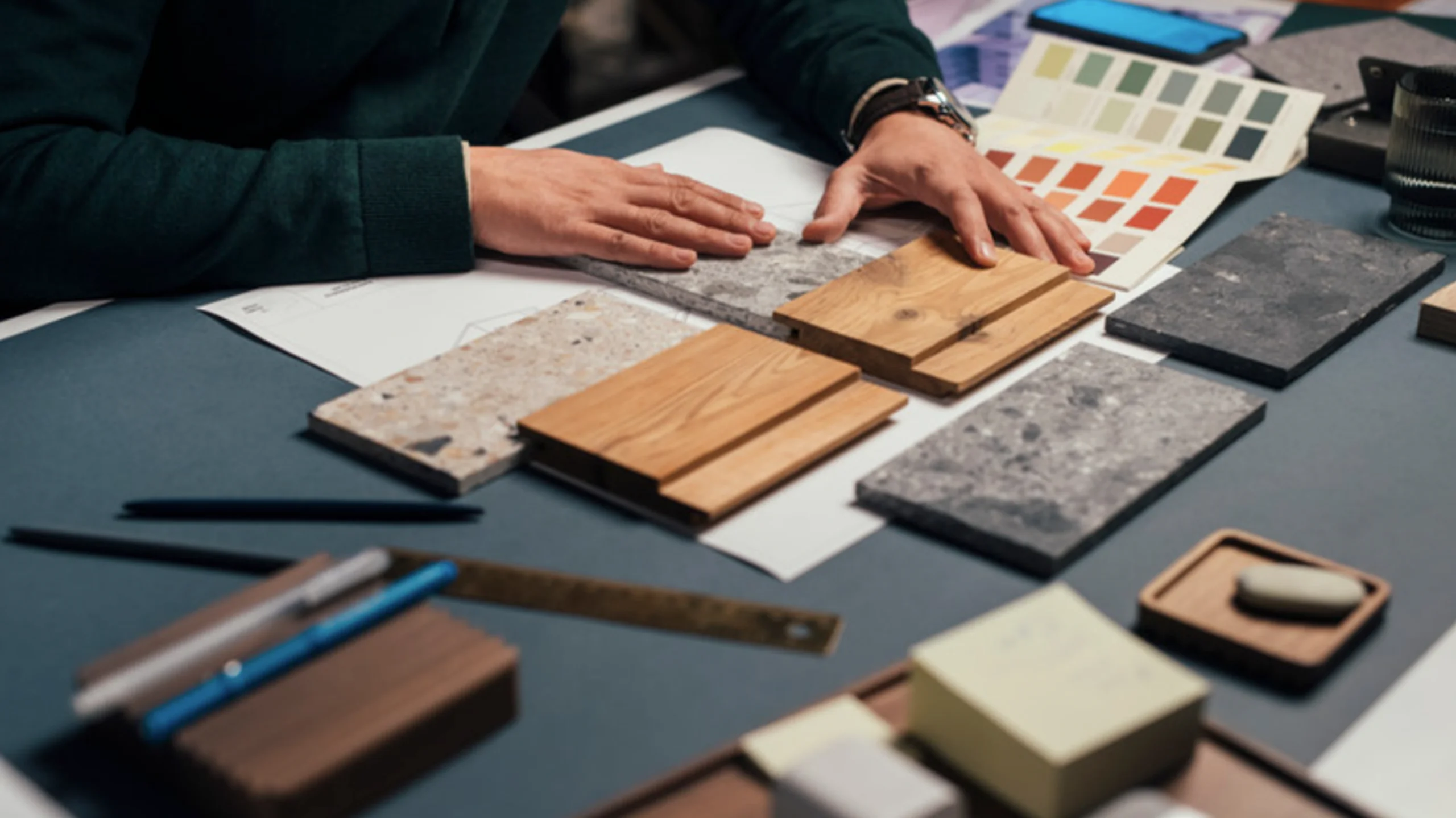 Hands arranging wood and stone material samples on a workspace with color swatch cards, design documents, pens, and a metal ruler