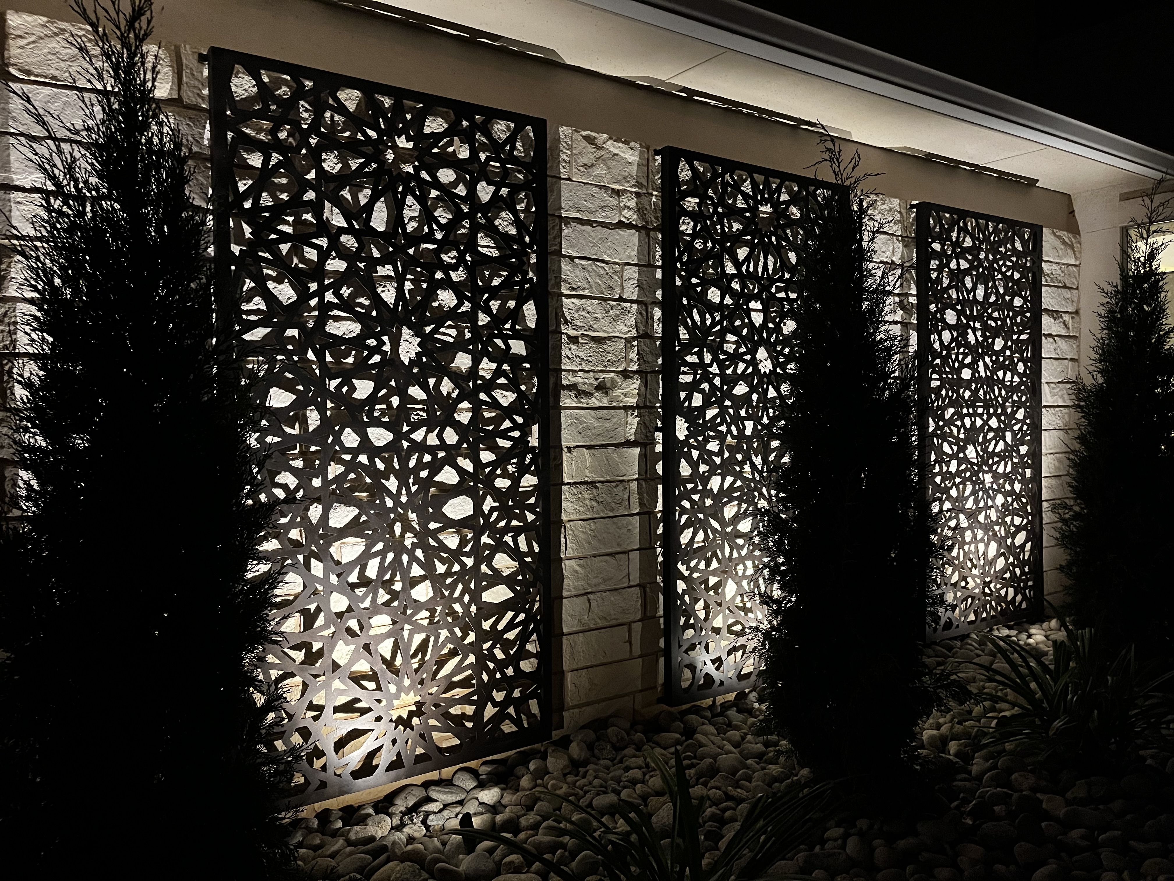 Three decorative metal screens with intricate cutout patterns mounted against a light stone exterior wall at night