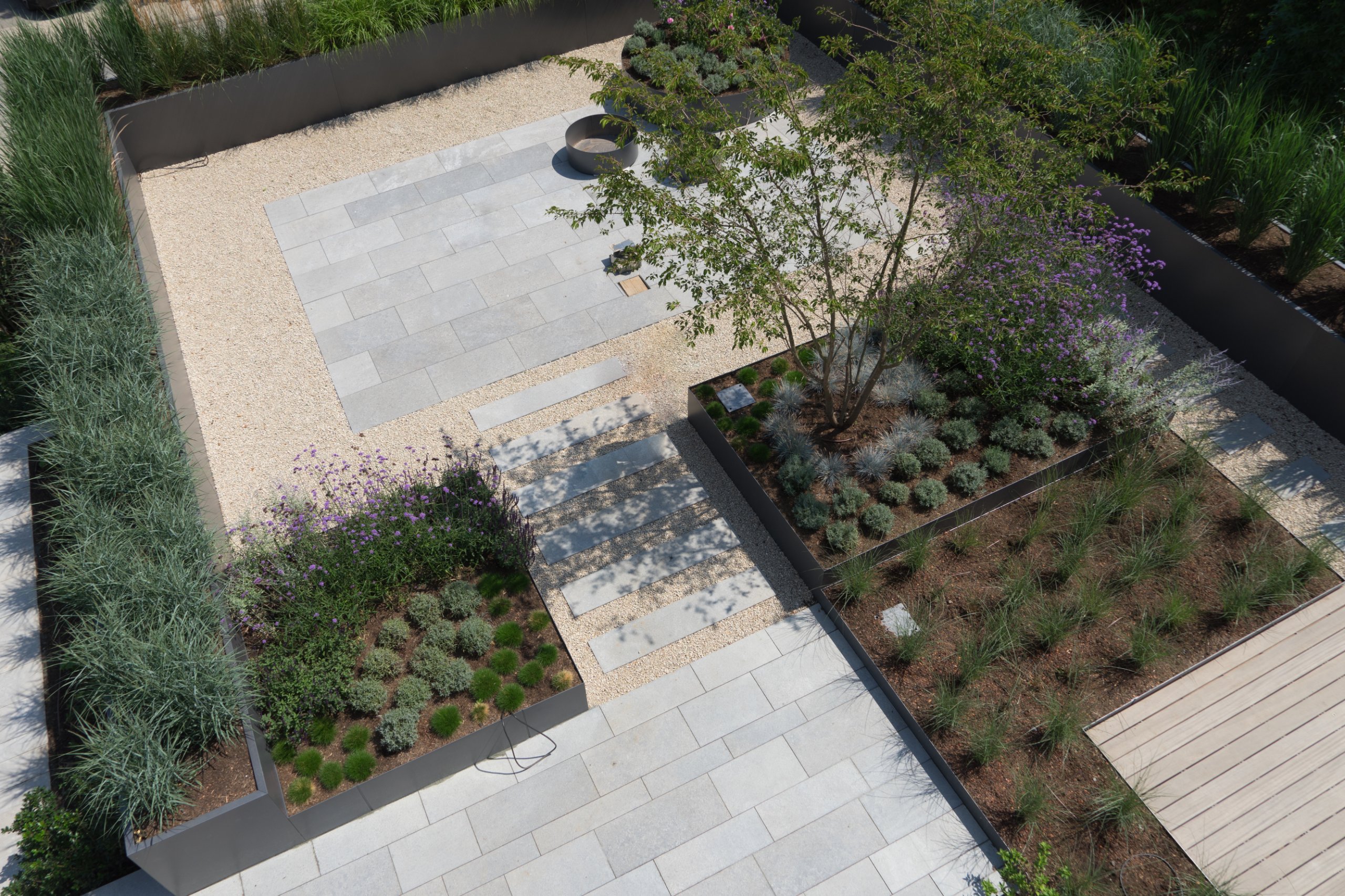 Overhead view of a modern landscaped courtyard with raised dark planters, ornamental grasses, purple flowers, grey stone pavers, a gravel stepping path, and a wooden deck area
