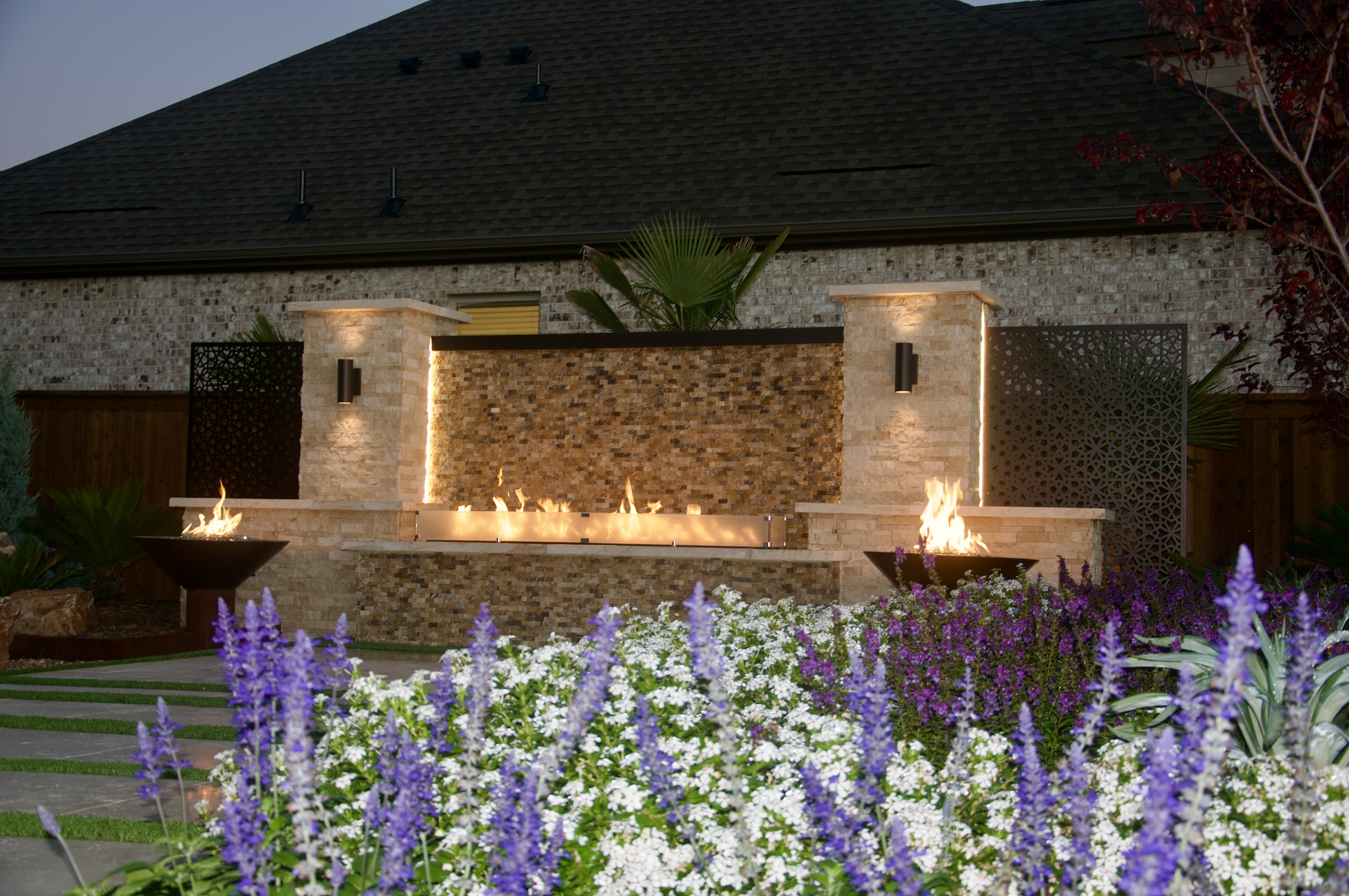Stone outdoor living structure with a central linear fire pit flanked by two lit fire bowls at dusk, in front of a brick home with decorative metal screens